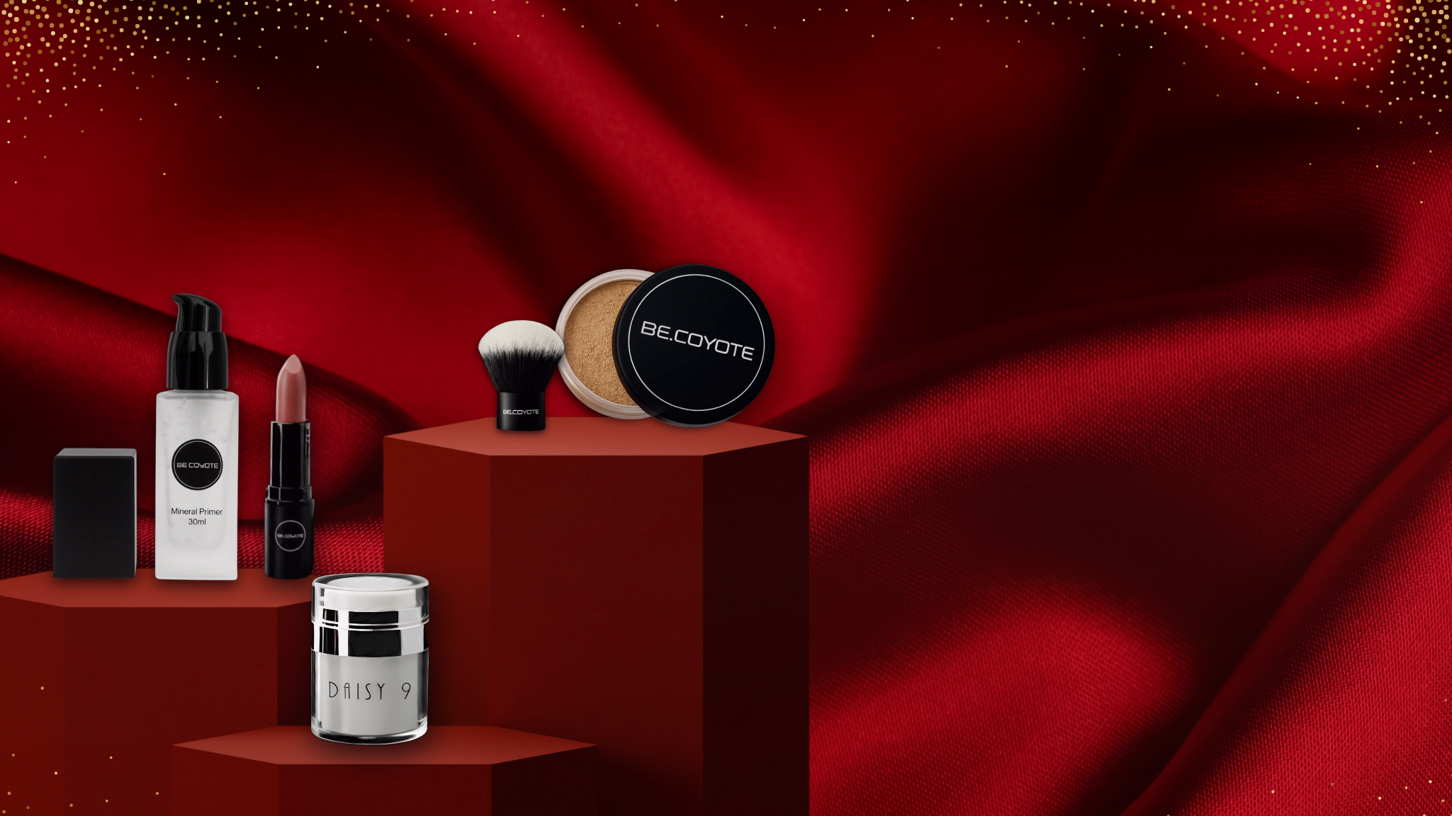 Be Coyote vegan, skin-kind makeup products displayed on deep red fabric with soft gold accents. Featured items include a mineral primer, ydrating lipstick, loose mineral foundation with kabuki brush, and Daisy 9 moisturiser, all arranged on red podium blocks.